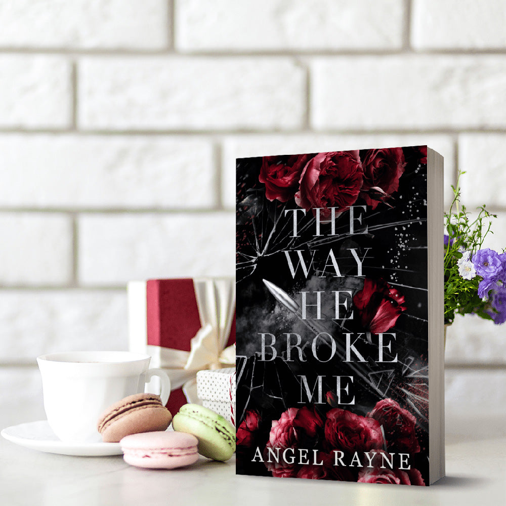 Book titled 'The Way He Broke Me' by Angel Rayne on a table with macarons and a cup.