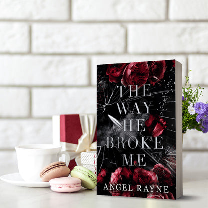 Book titled 'The Way He Broke Me' by Angel Rayne on a table with macarons and a cup.
